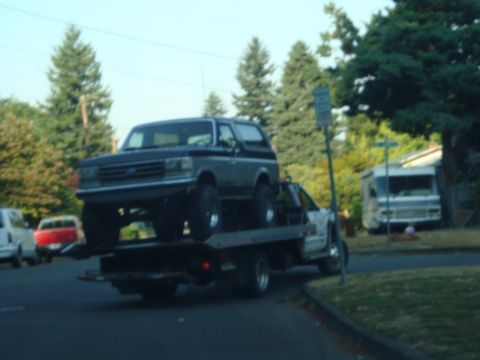 1989 Ford Bronco - A dark SUV with a roof rack is parked on the side of a street, with trees and other vehicles visible.