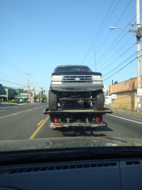 1989 Ford Bronco - Rear view of a dark SUV driving on a highway under a clear blue sky.