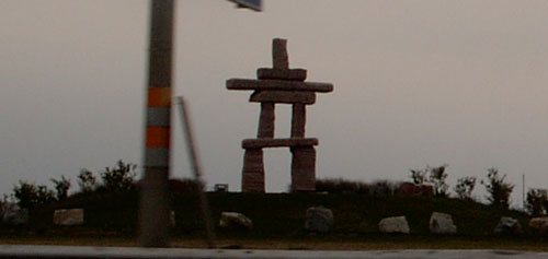 Inukshuk statue on a grassy hill with a utility pole in the foreground. Supermotors.net logo in the corner
