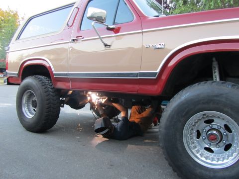 1984 Ford Bronco - A mechanic works on the undercarriage of a lifted Ford Bronco, sparks flying.
