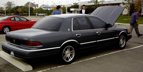 1999 Ford Crown Victoria - A dark sedan, likely a Mercury Grand Marquis, is parked with its hood open, while another red car is visible in the background.