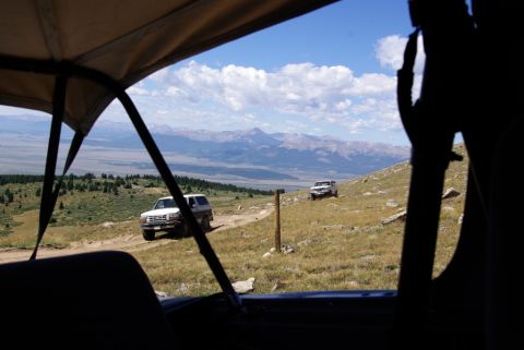 1996 Ford Bronco - Two SUVs traverse a mountain trail with a vast valley and mountain range in the background.