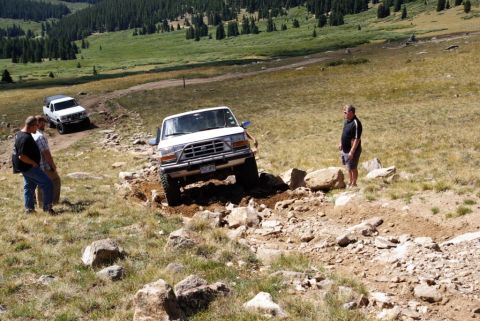 1996 Ford Bronco - A white Ford Bronco is shown off-roading on a rocky mountain trail with another vehicle in the background.