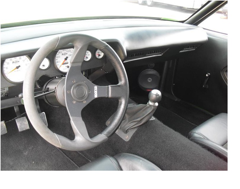 Interior view of a car with a large aftermarket steering wheel, custom gauges, and a manual shifter