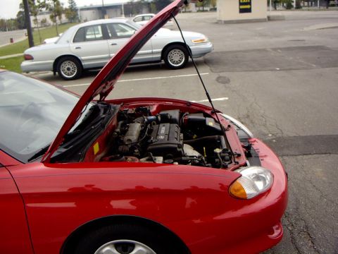 1999 Ford Crown Victoria - The engine bay of a red car is open, with a silver sedan parked behind it.