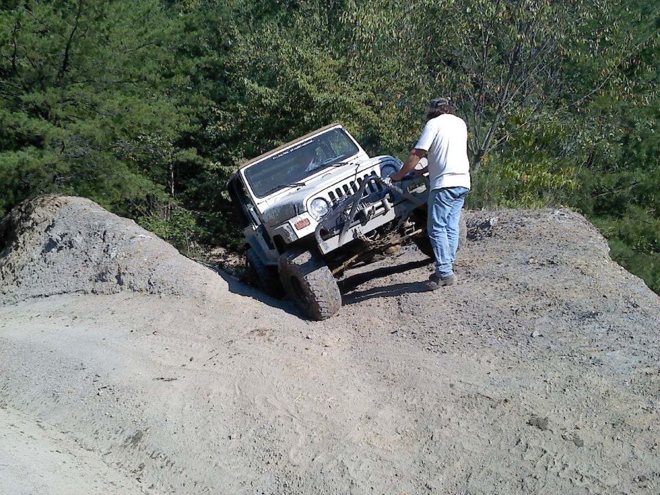 White Jeep Wrangler TJ tilted on a dirt incline, man in white shirt and jeans standing by the front