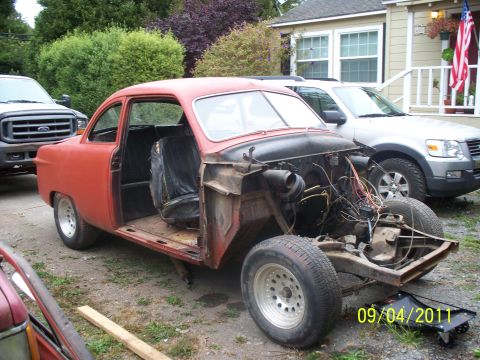 1990 Ford Bronco - This red coupe is missing its engine and interior components, parked in a driveway with other vehicles.