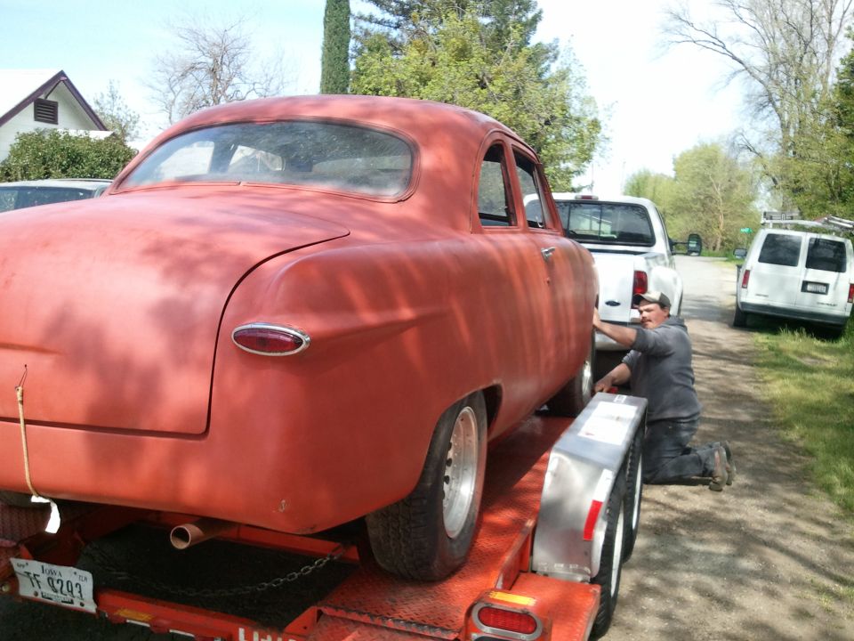 Red sedan on trailer, man securing it, white pickup and van in background