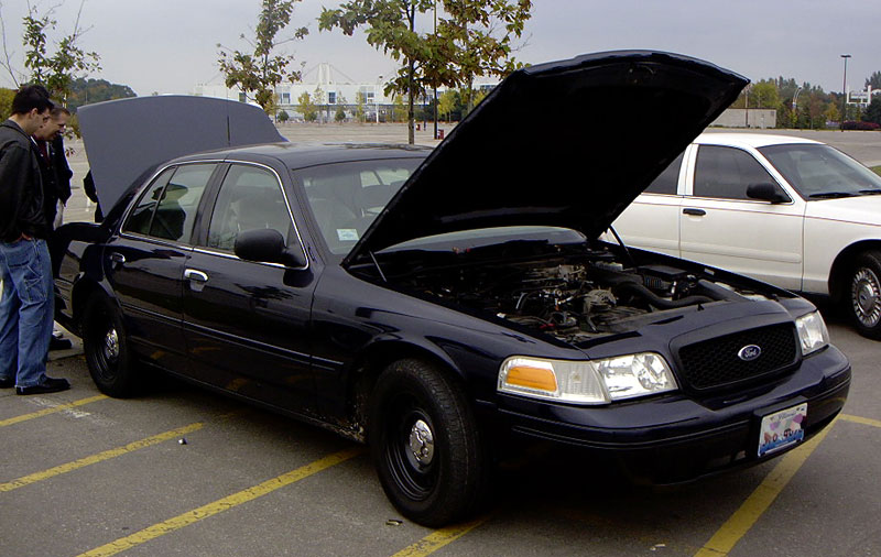 Dark blue Ford Crown Victoria with hood open, showing engine. White Crown Victoria parked behind. Two people looking at the dark blue car