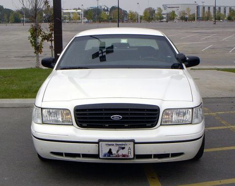1999 Ford Crown Victoria - A white 1999 Ford Crown Victoria is parked in a large, empty parking lot with buildings in the background.