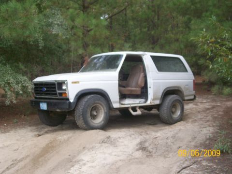 1983 Ford Bronco - White Ford Bronco parked on a dirt path, showing its interior and off-road tires.