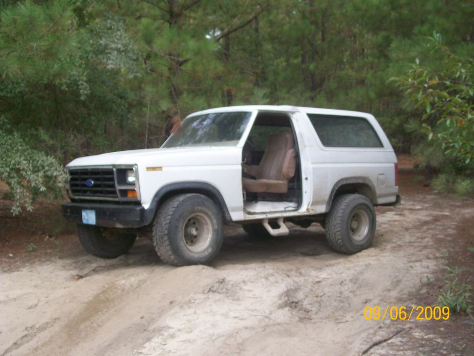 White Ford Bronco with driver's door open, parked on a dirt path with trees in the background
