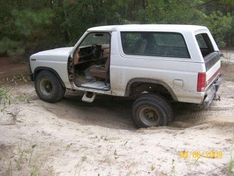 1983 Ford Bronco - This white Ford Bronco is stuck in the sand, with its driver's door open and the front right wheel partially submerged.