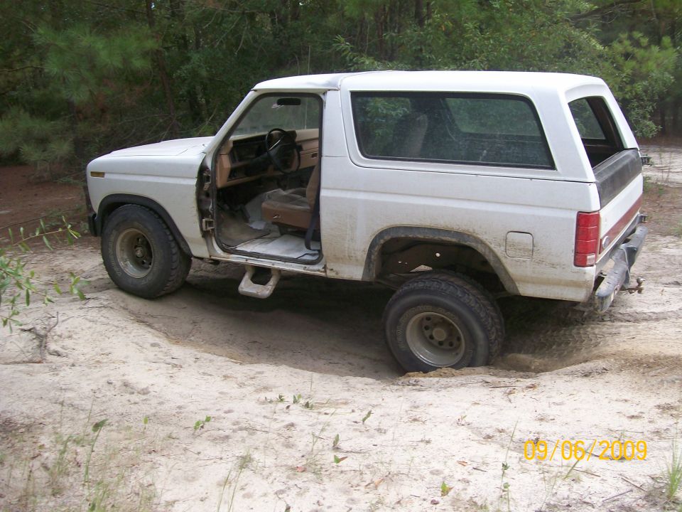 White 1980s Ford Bronco with driver's door open, stuck in sand with front right wheel buried