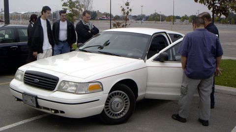 1999 Ford Crown Victoria - A group of people inspect a white 1999 Ford Crown Victoria, its front passenger door ajar.
