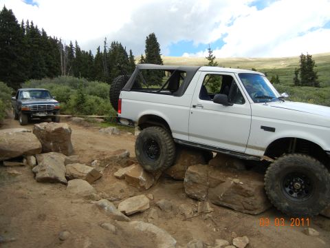1996 Ford Bronco - A white 1996 Ford Bronco tackles a rocky off-road trail, with a dark-colored truck assisting in the background.