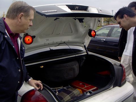 1999 Ford Crown Victoria - The trunk of a white car is open, revealing a spare tire and some items inside. Another car is visible in the background.