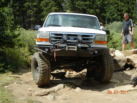 1996 Ford Bronco - A white 1996 Ford Bronco navigates a challenging rocky terrain, equipped with a winch and off-road tires.