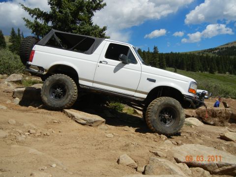 1996 Ford Bronco - A white Ford Bronco navigates a challenging rocky terrain under a blue sky with scattered clouds.