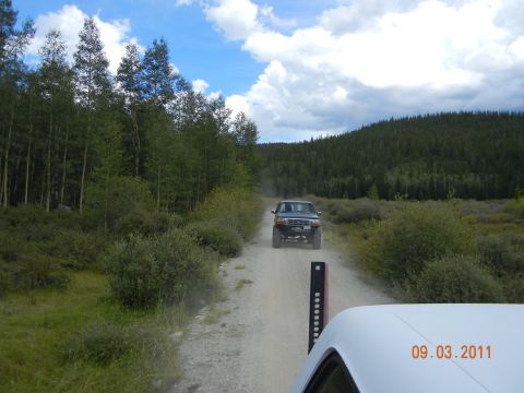 1996 Ford Bronco - A dark green Ford Bronco drives down a dusty dirt road surrounded by trees and hills.