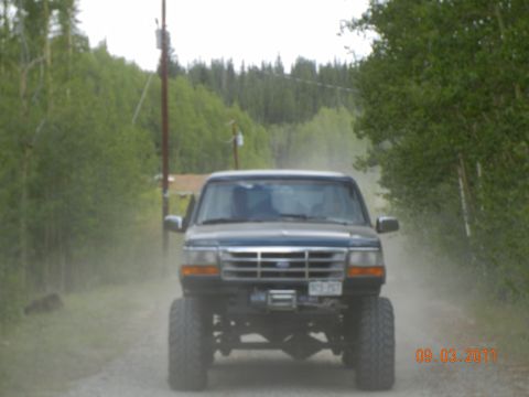 1996 Ford Bronco - This lifted Ford pickup truck is driving down a dusty dirt road surrounded by trees.