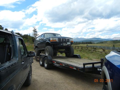 1996 Ford Bronco - A lifted Ford Bronco is transported on a trailer, with another vehicle in the foreground and mountains in the background.