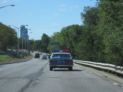 1991 Ford LTD Crown Victoria - blue police sedan marked EMER 911 with roof lights drives ahead on a highway curving toward distant city towers.