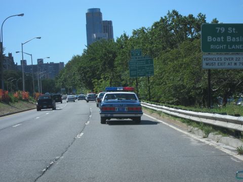 1991 Ford LTD Crown Victoria - blue police sedan with roof lights travels a tree-lined highway past a green 79 St Boat Basin overhead exit sign.