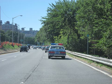 1991 Ford LTD Crown Victoria - blue police sedan with a roof lightbar leads traffic down a tree-lined parkway with city buildings on the horizon.