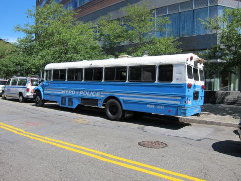 1991 Ford LTD Crown Victoria - blue and white NYPD Police bus marked PBMS 3328 is parked curbside next to a modern glass building and trees.