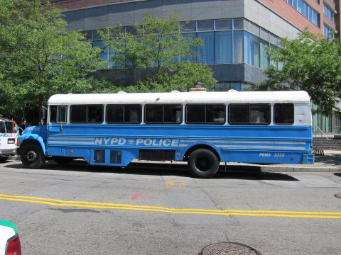 1991 Ford LTD Crown Victoria - Side view of a blue and white NYPD Police bus lettered PBMS 3328 parked along a street in front of an office building.