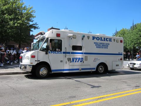 1991 Ford LTD Crown Victoria - white NYPD Police Mobile Command Center truck is parked curbside as kilted bagpipers stand on the sidewalk behind it.