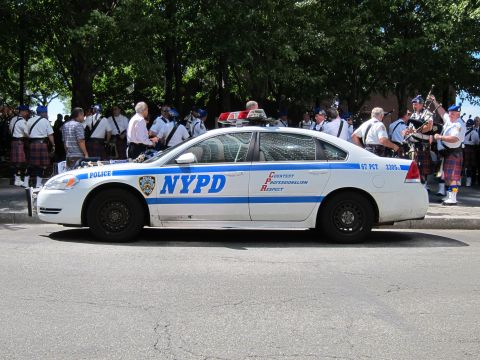 1991 Ford LTD Crown Victoria - white NYPD patrol sedan is parked on a street while officers and kilted bagpipers gather along the sidewalk behind it.