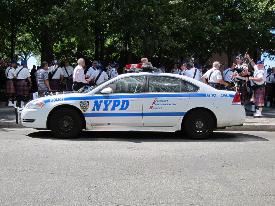 White NYPD police sedan parked on a street with police officers and kilted bagpipers behind