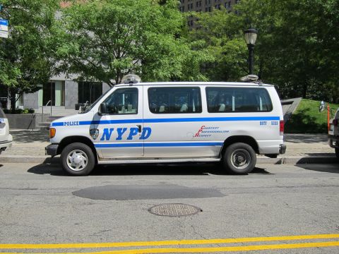 1991 Ford LTD Crown Victoria - white NYPD police passenger van with a blue stripe and roof lights is parked along a curb on a sunny city street.