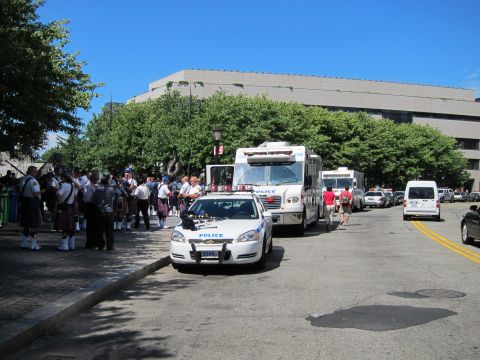 1991 Ford LTD Crown Victoria - white Police patrol car and command bus parked on a sunny city street as a bagpipe band in kilts gathers on the sidewalk.