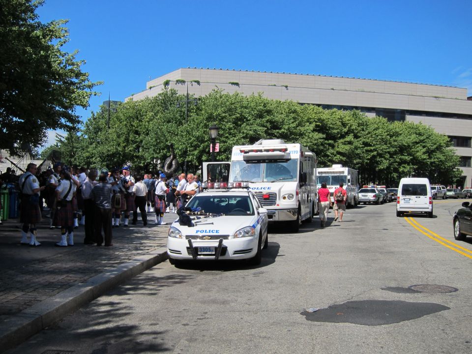 White police car and bus on city street with bagpipe band in kilts nearby