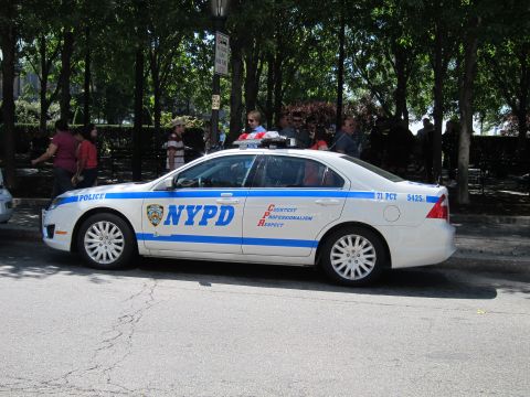 1991 Ford LTD Crown Victoria - white NYPD patrol sedan with blue striping and CPR lettering, parked beside a tree-lined park fence.