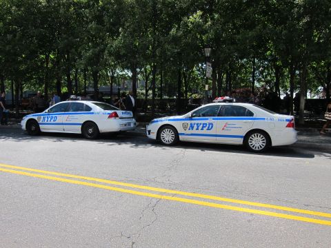 1991 Ford LTD Crown Victoria - Two white-and-blue NYPD patrol sedans parked nose-to-tail along a tree-lined city street.