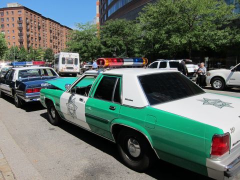 1991 Ford LTD Crown Victoria - vintage green-and-white police sedan with a star emblem parked next to a blue emergency unit on a city street.