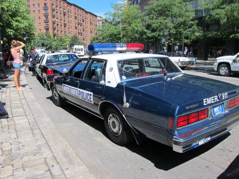 1991 Ford LTD Crown Victoria - dark blue vintage Chevrolet police sedan marked EMER 911 parked along a cobblestone curb on a sunny street.