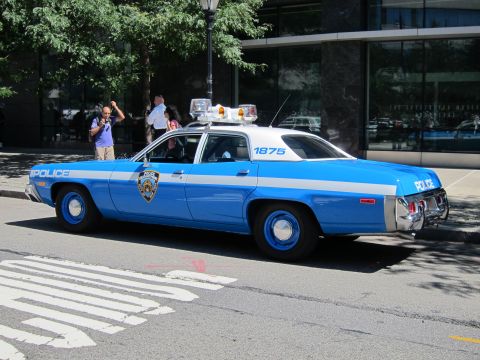 1991 Ford LTD Crown Victoria - vintage blue-and-white NYPD police sedan, unit 1875, parked over a crosswalk on a sunny city street.