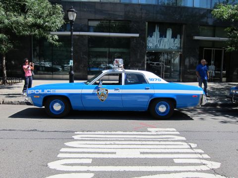 1991 Ford LTD Crown Victoria - Side profile of a vintage blue-and-white NYPD police sedan, unit 1875, parked over a crosswalk near a glass building.