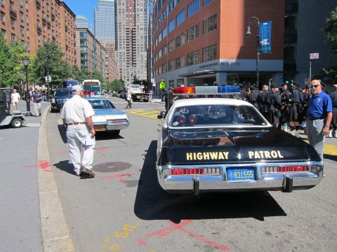 1991 Ford LTD Crown Victoria - Rear view of a black Highway Patrol sedan parked on a city street as a man in white walks past and crowds gather.