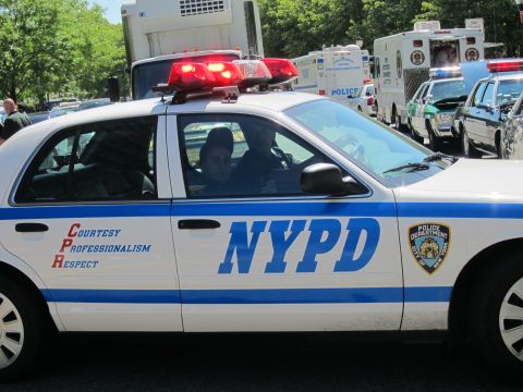 1991 Ford LTD Crown Victoria - white-and-blue NYPD patrol car with roof lights activated amid a line of emergency vehicles on a tree-lined street.