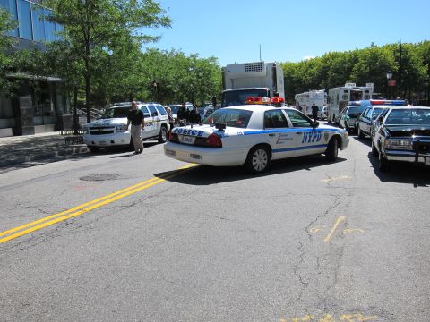 1991 Ford LTD Crown Victoria - white-and-blue NYPD patrol car turning on a wide city street lined with police vans, SUVs and a bus.