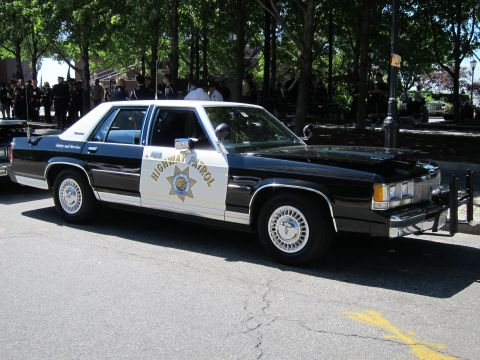 1991 Ford LTD Crown Victoria - black-and-white vintage Ford Highway Patrol sedan with a star emblem parked along a tree-lined curb.