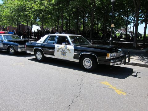 1991 Ford LTD Crown Victoria - Black-and-white vintage Highway Patrol sedans parked in a row along a tree-lined street with a crowd in the background.