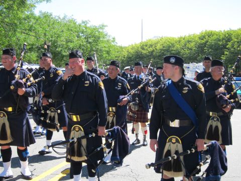 1991 Ford LTD Crown Victoria - Members of a police pipe band in tartan kilts and dark uniforms carry bagpipes while marching outdoors on a sunny day.