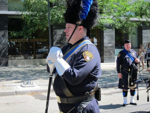 1991 Ford LTD Crown Victoria - bagpiper in a dark police uniform and tall feathered bearskin hat stands on a city sidewalk, with another piper behind him.